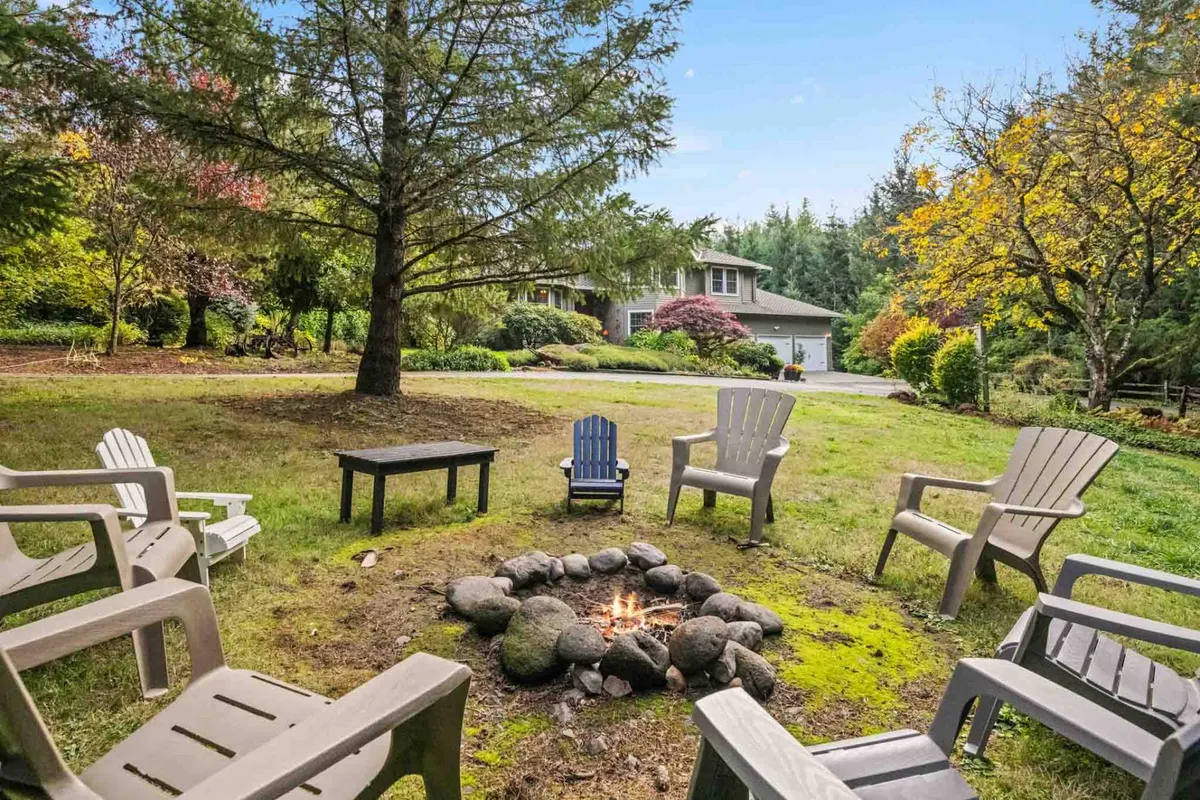 An inviting fire pit is encircled by comfortable chairs beneath a leafy tree canopy, with the house softly illuminated in the background. The setup is ideal for evening gatherings, storytelling, or simply savoring the warmth of the fire under the stars.