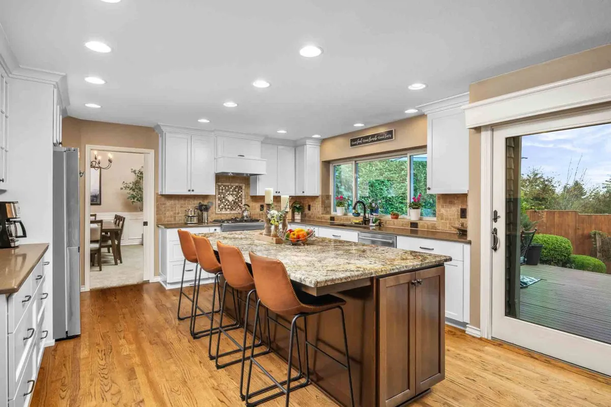 A stylish, fully equipped kitchen with granite countertops and a central island, set against a backdrop of large windows framing outdoor greenery. The warm, open layout invites both cooking and conversation, seamlessly connecting to the living and dining areas.