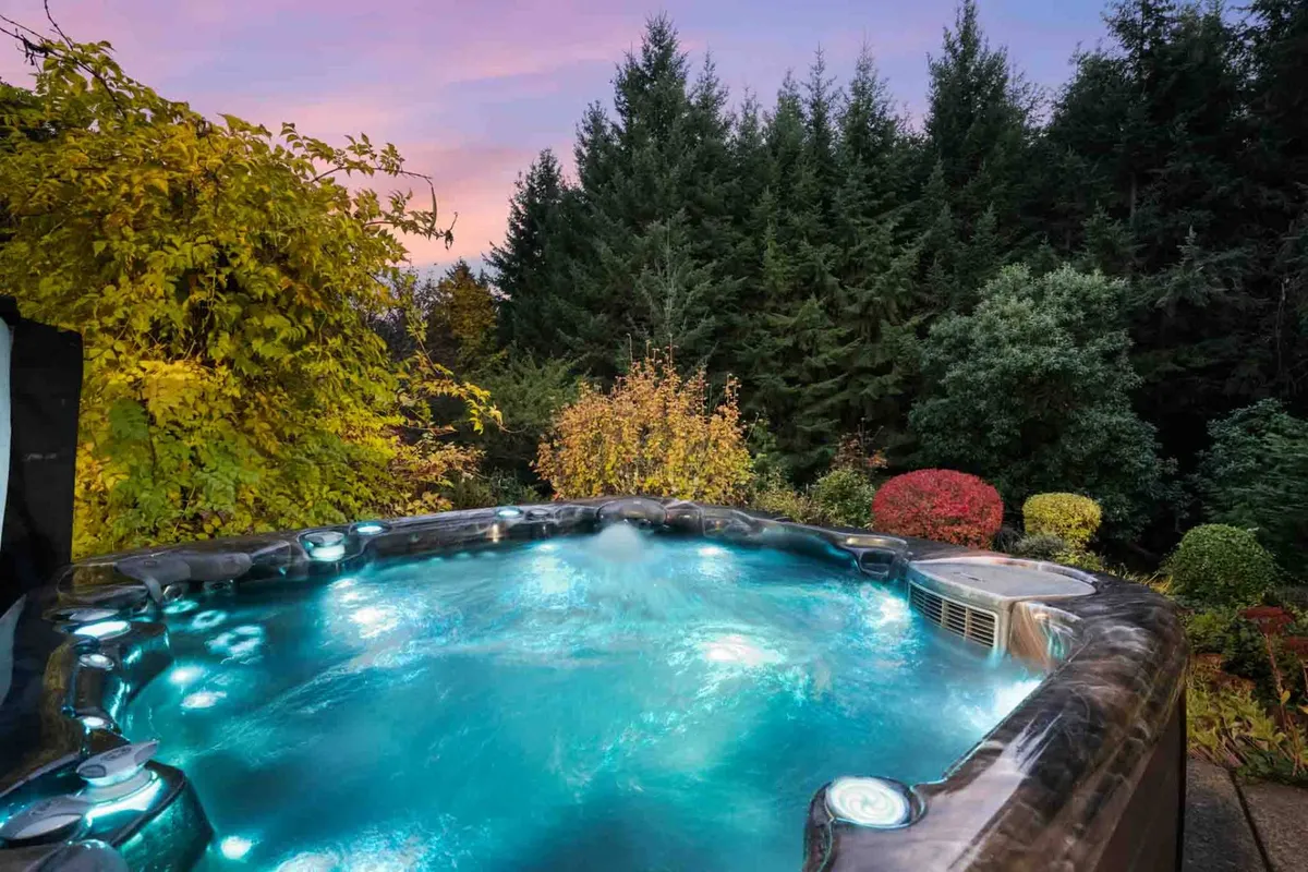 This exterior image features a hot tub surrounded by lush foliage and distant trees. The setting is tranquil, with the soft evening light suggesting a perfect spot for relaxing or stargazing after a day out. The hot tub is the focal point, inviting groups to socialize and unwind in privacy and comfort.