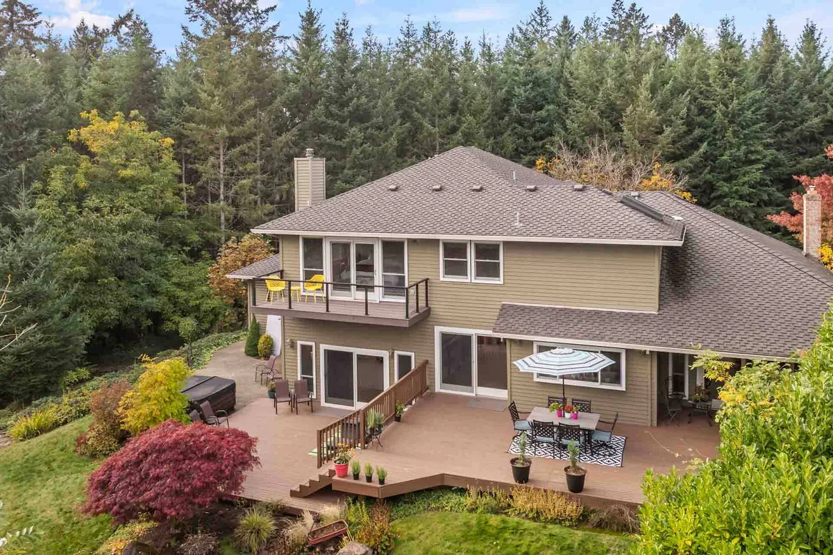 A spacious deck stretches out from the home, furnished with colorful outdoor seating and surrounded by lush greenery. The firepit area is visible to one side, and large windows reflect the tranquil landscape. The overall mood is inviting, ideal for outdoor dining, family gatherings, and sunset watching.