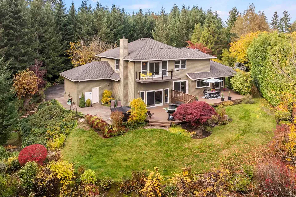 A serene exterior view of the house nestled amid mature trees, with a spacious deck overlooking a colorful garden. The surrounding landscape exudes tranquility and privacy, with lush greenery creating a sense of seclusion. The deck area is the focal point, inviting relaxation or al fresco dining while immersed in nature.
